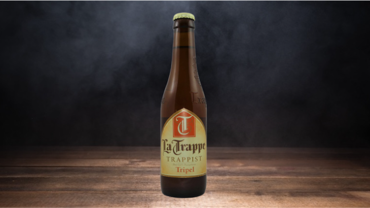 Braune Glasflasche des Bieres „La Trappe Trappist Tripel“ mit gelbem Etikett und roter Schrift auf Holztisch vor dunklem Hintergrund.