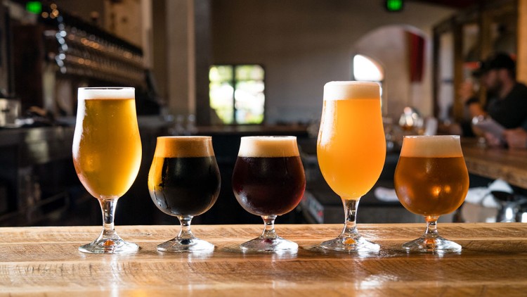 Fünf gefüllte Biergläser in verschiedenen Größen und Farben stehen auf einem Holzcounter in einer Bar mit unscharfem Hintergrund.