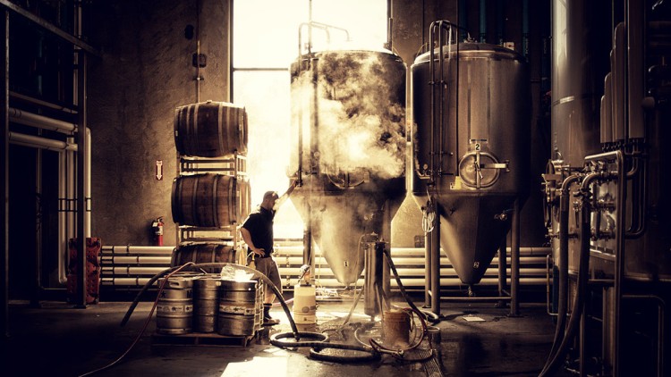 Person in Arbeitskleidung steht in einer Brauerei neben silbernen Tanks, Holzfässern und Schläuchen, aus einem Tank steigt Dampf.