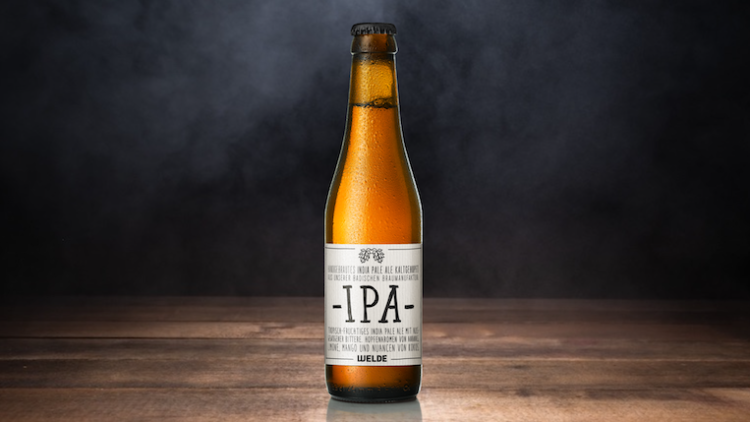 Braunglasflasche mit weißem Etikett, großem schwarzen Schriftzug „IPA“ und kleinem Text darunter auf einem Holztisch vor dunklem Hintergrund.