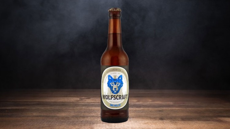 Braune Bierflasche mit Etikett und blauem Wolfskopf auf Holzuntergrund vor dunklem, rauchigem Hintergrund.