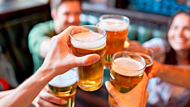 Mehrere Hände stoßen mit gefüllten Biergläsern in einem gemütlichen Raum mit Holzmöbeln an.