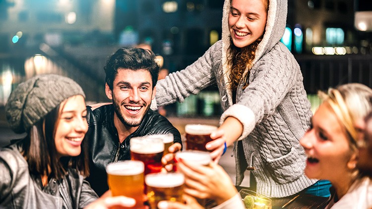 Menschen heben gefüllte Biergläser an einem Holztisch in stimmungsvoller Außenbeleuchtung zum Anstoßen.