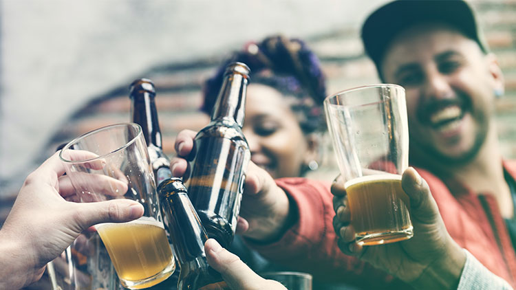 Mehrere Hände stoßen mit Bierflaschen und Gläsern mit Bier vor unscharfem Hintergrund an.