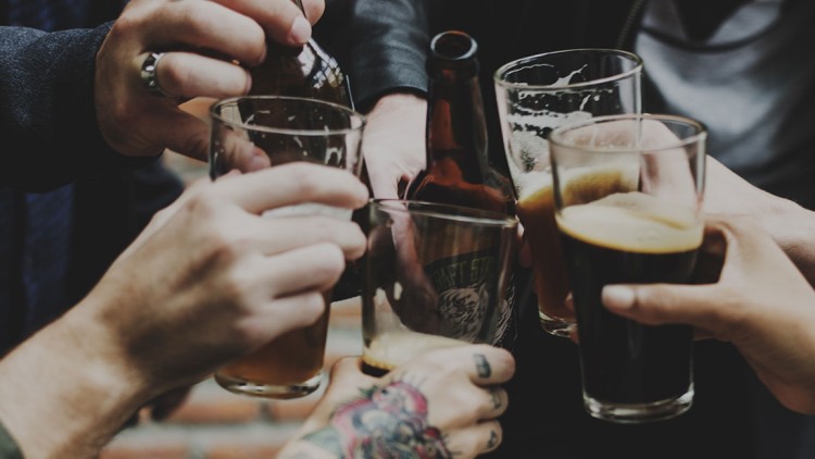 Mehrere Hände stoßen mit Gläsern und einer Bierflasche an, im Hintergrund unscharfe Kleidung und eine Backsteinmauer.