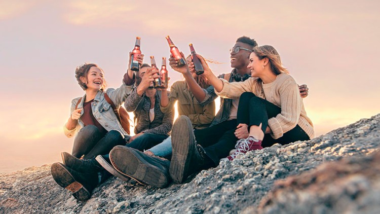 Mehrere Personen sitzen auf einem Felsen vor Sonnenuntergang und stoßen mit Flaschen an.