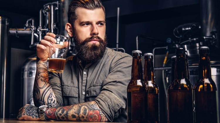 Person in olivgrünem Hemd mit tätowierten Armen hält ein Glas Bier neben mehreren braunen Bierflaschen in einer Brauerei.