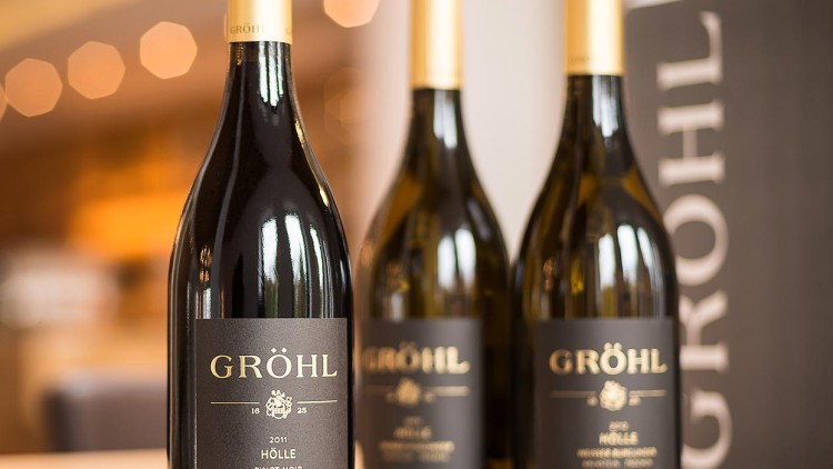 Drei Weinflaschen mit schwarzem Etikett und goldener Aufschrift „GRÖHL“, im Hintergrund unscharfe Beleuchtung.