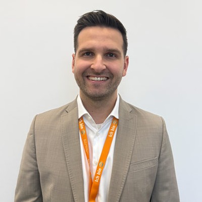 Person in beigem Sakko mit weißem Hemd trägt ein orangefarbenes „ICH BIN GLOBUS“-Lanyard vor weißem Hintergrund.