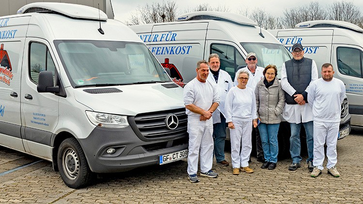 Sechs Personen in Arbeitskleidung stehen vor drei weißen Lieferwagen mit der Aufschrift „Natur für Feinkost“.
