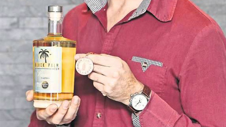 Person in rotem Hemd hält eine Flasche „Black Palm Rum“ mit gelbem Etikett und eine runde Medaille vor einer grauen Wand.