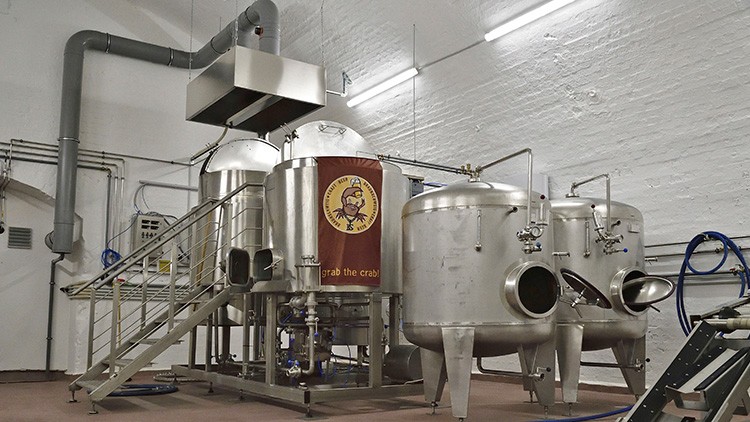 Mehrere Edelstahl-Braukessel mit Treppe, Rohren und einem Logo mit einem roten Schriftzug „grab the crab!“ in einer Brauerei.