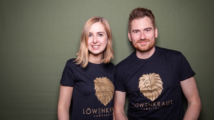 Zwei Personen in schwarzen T-Shirts mit goldenen Löwenmotiven und der Aufschrift „LÖWENKRAUT GEWÜRZREIHE“ vor grünem Hintergrund.