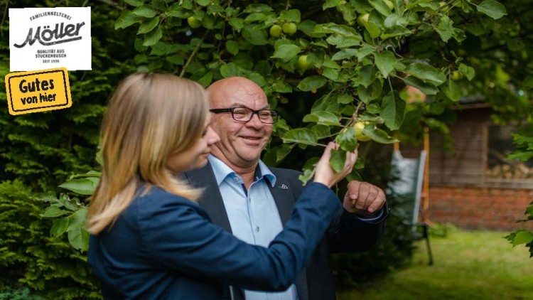 Zwei Personen inspizieren einen Ast mit grünen Äpfeln, im Hintergrund ist ein hölzernes Gebäude und Bäume zu sehen.