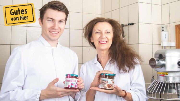 Zwei Personen in weißen Kitteln halten Gläser mit Himbeerkonfitüre und Schokocreme, links oben Schriftzug „Gutes von hier“.