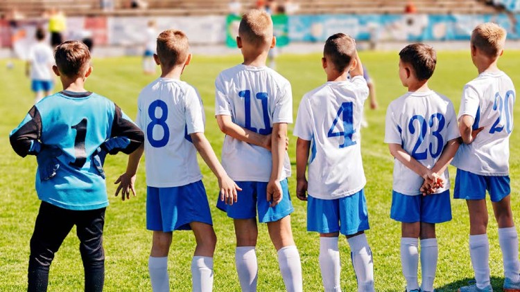 Sechs Kinder in weißen Trikots mit blauen Details stehen auf einem Fußballfeld, Rückennummern sind sichtbar.