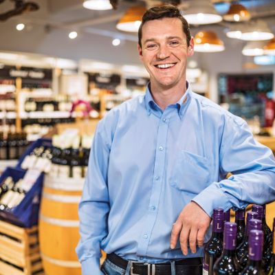 Eine Person in hellblauem Hemd und Jeans steht in einem Weinregalbereich mit Flaschen und einer Holzfass-Dekoration.