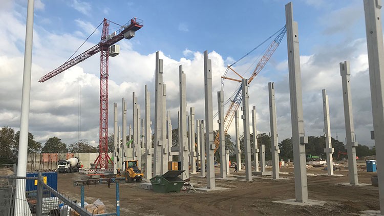 Mehrere Betonpfeiler stehen auf einer Baustelle, umgeben von zwei Kränen, Baugeräten und wolkigem Himmel.