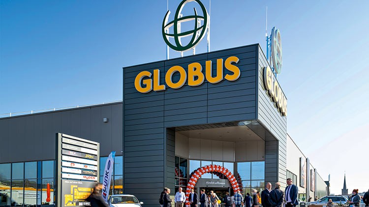 Eingangsbereich eines GLOBUS-Markts mit rotem und weißem Luftballonbogen, mehreren Menschen und sichtbarem GLOBUS-Logo.