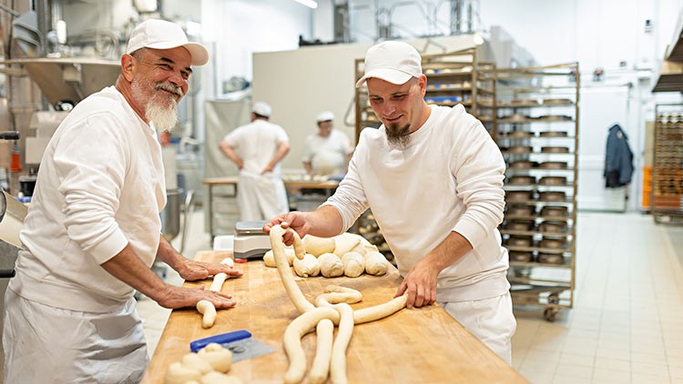 Personen in weißer Arbeitskleidung formen Teigstränge an einem Holztisch in einer Backstube mit Regalen im Hintergrund.