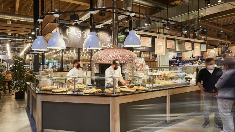 Pizzastand mit Holzofen, Glasvitrine mit belegten Pizzen, Schriftzug Pizza im Hintergrund, helle Lampen hängen von der Decke.