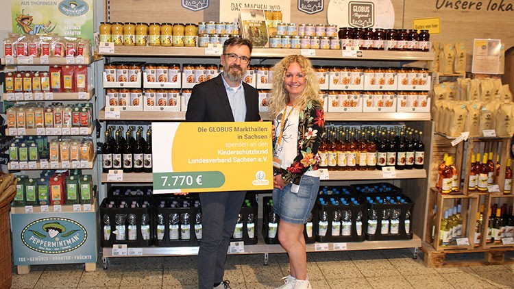 Zwei Personen halten einen gelben Spendenscheck mit dem Betrag 7.770 Euro vor einem Regal mit Marmeladen und Getränken.