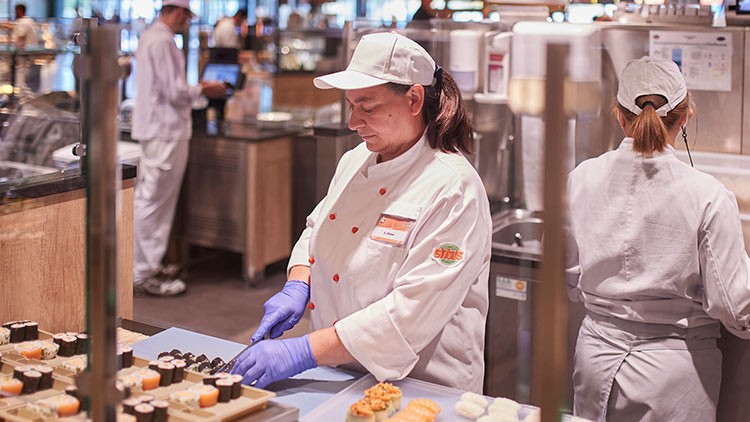 Eine Person in weißer Kochkleidung und blauen Handschuhen bereitet Sushi in einer GLOBUS-Sushi-Theke zu.