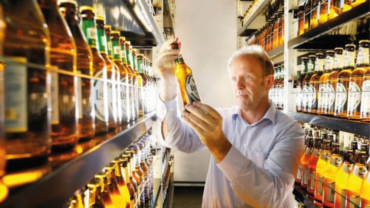 Person betrachtet eine Bierflasche auf einem gut beleuchteten Gang mit Regalen voller weiterer Flaschen in einem Getränkemarkt.