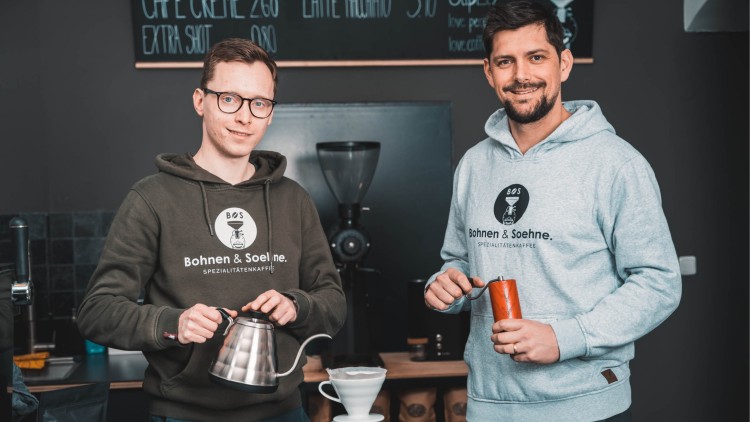 Zwei Personen mit Bohnen & Soehne-Pullovern stehen vor einer Kaffeemühle mit Handfilterkanne und Kaffeemühle in den Händen.