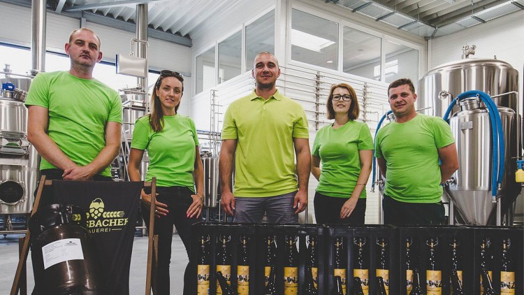 Fünf Personen in grünen T-Shirts stehen vor Brauereigeräten und gestapelten Bierflaschen mit gelbem Etikett.