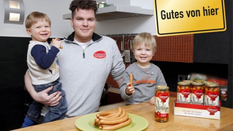 Auf einem Tisch stehen Würstchen auf einem grünen Teller sowie Gläser mit der Aufschrift „Eidmann Frankfurter“, daneben ein gelbes Schild mit „Gutes von hier“.