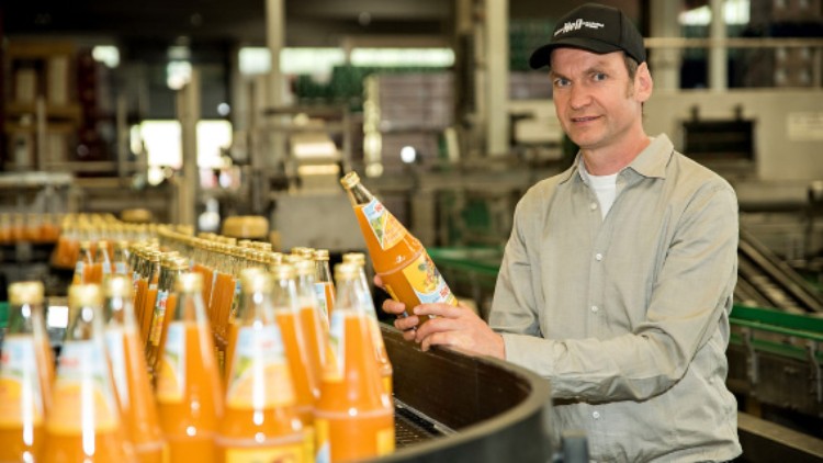 Person hält eine Flasche mit orangener Flüssigkeit an einem Fließband mit weiteren identischen Flaschen in einer Fabrikhalle.