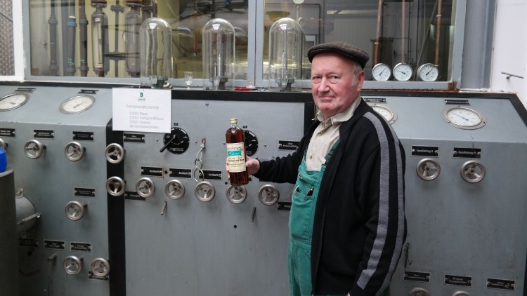 Person in grüner Arbeitskleidung hält eine Flasche vor einer grauen Schalttafel mit Druckanzeigen und Beschriftungen.