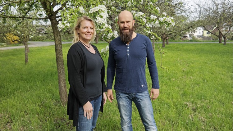 Zwei Personen stehen auf einer grünen Wiese vor blühenden Obstbäumen, im Hintergrund sind Häuser und eine Straße.