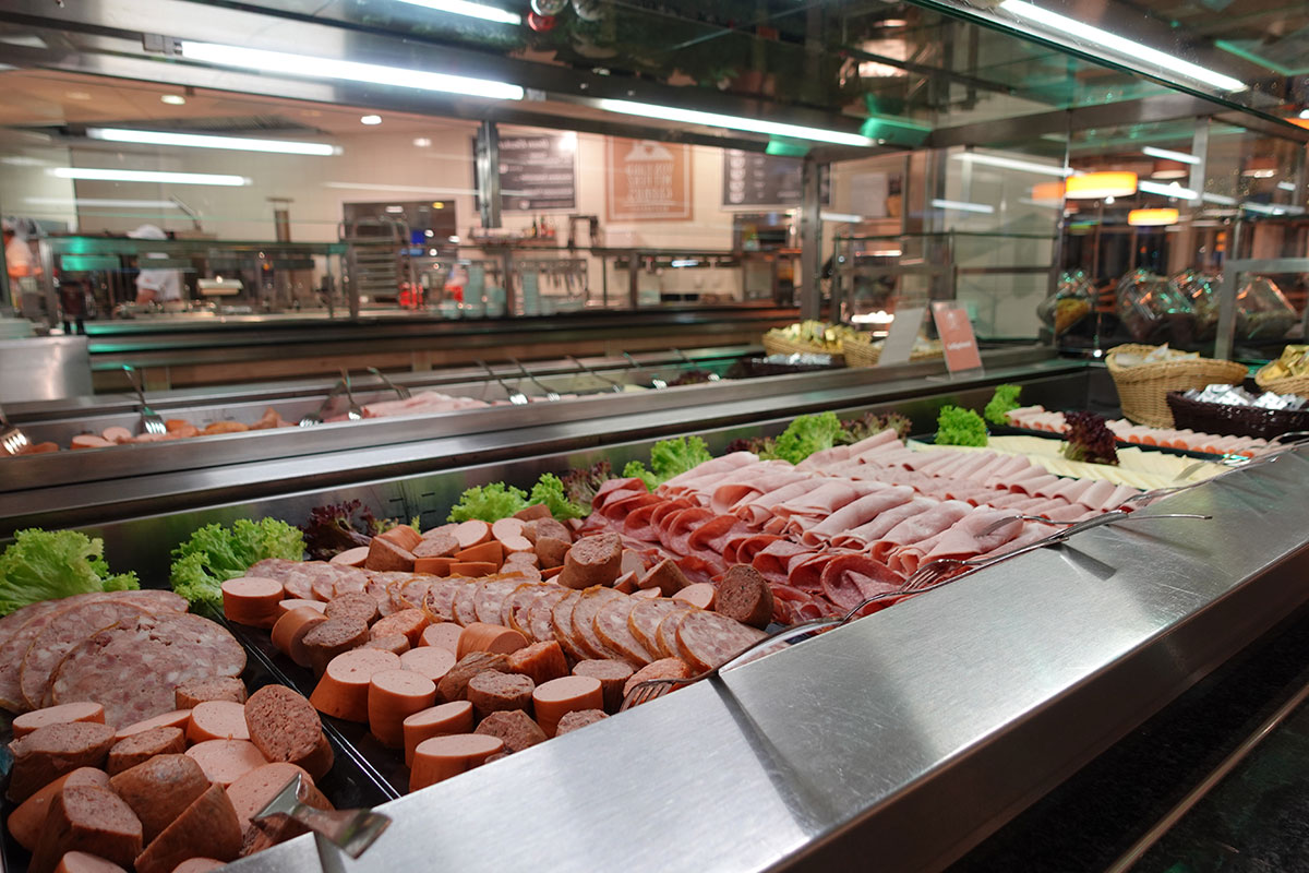 Theke mit Auswahl an geschnittenen Wurstsorten, belegten Brötchen, frischen Salaten und Tellern in einem hellen Verkaufsraum.