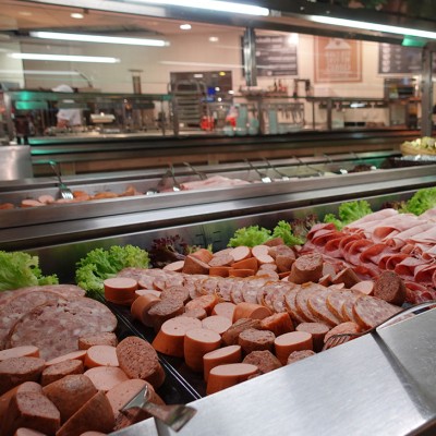 Theke mit Auswahl an geschnittenen Wurstsorten, belegten Brötchen, frischen Salaten und Tellern in einem hellen Verkaufsraum.