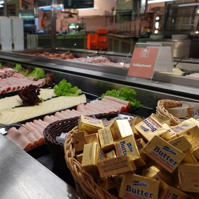 Eine Theke mit Geflügelwurst, Käse, Salat, einem Schild mit der Aufschrift „Geflügelwurst“ und Körben mit Butterstücken.