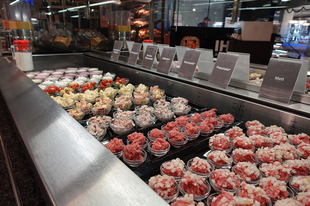 Frische Theke mit kleinen Schalen verschiedener Fleisch- und Wurstsalate, Mett und beschrifteten Schildern auf Edelstahl.