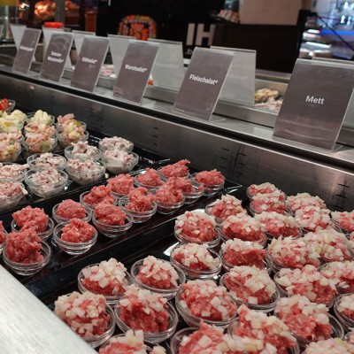 Frische Theke mit kleinen Schalen verschiedener Fleisch- und Wurstsalate, Mett und beschrifteten Schildern auf Edelstahl.