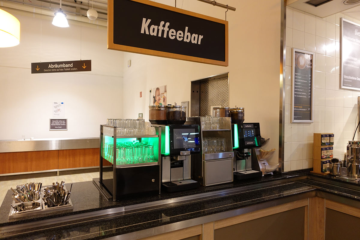 Kaffeebar mit zwei Kaffeemaschinen, leeren Gläsern, Besteck, einem Schild „Abräumband“ und weiteren Hinweistafeln.