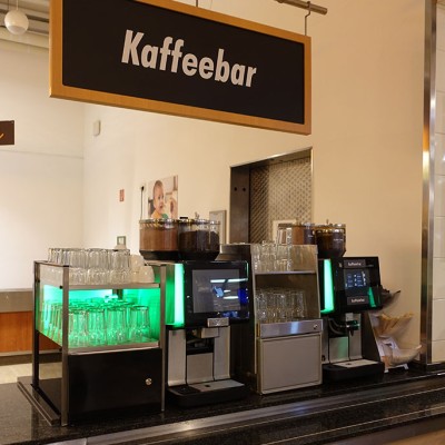Kaffeebar mit zwei Kaffeemaschinen, leeren Gläsern, Besteck, einem Schild „Abräumband“ und weiteren Hinweistafeln.