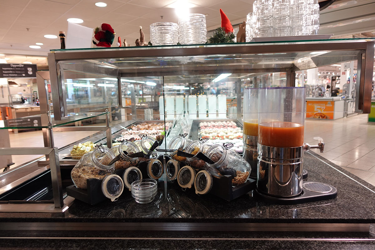 Glasbehälter mit Müsli und Spender für Saft auf einer Theke vor einer Auswahl an Desserts in einem Gastronomiebereich.