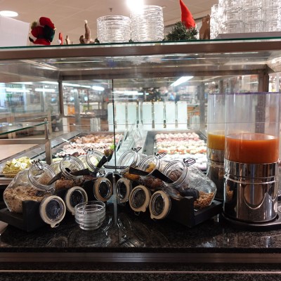 Glasbehälter mit Müsli und Spender für Saft auf einer Theke vor einer Auswahl an Desserts in einem Gastronomiebereich.