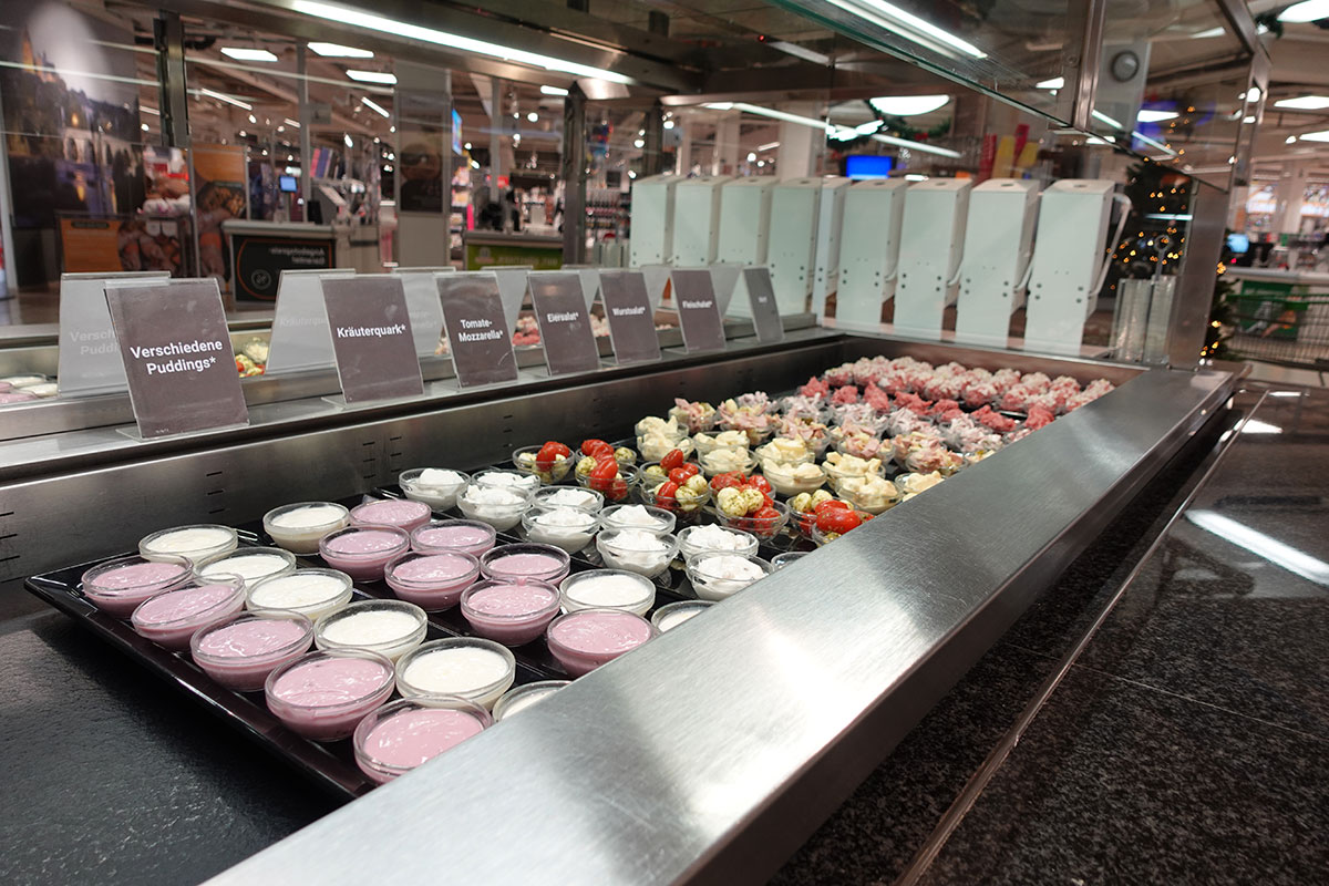 Offene Kühltheke mit kleinen Schalen verschiedener Speisen und Beschilderungen wie „Verschiedene Puddings“ und „Kräuterquark“.