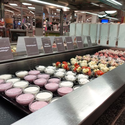 Offene Kühltheke mit kleinen Schalen verschiedener Speisen und Beschilderungen wie „Verschiedene Puddings“ und „Kräuterquark“.