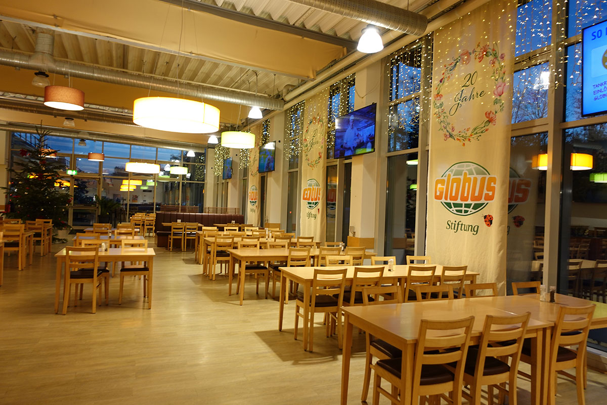 Ein heller Speiseraum mit Holzstühlen und Tischen, dekoriert mit Bannern der Globus-Stiftung und einem Weihnachtsbaum.