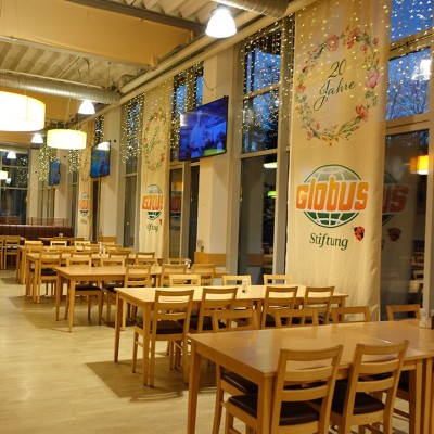 Ein heller Speiseraum mit Holzstühlen und Tischen, dekoriert mit Bannern der Globus-Stiftung und einem Weihnachtsbaum.