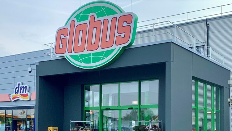 Eingangsbereich eines GLOBUS Markts mit grünem Logo darüber, Pflanzenständern und Zebrastreifen, daneben ein dm-Logo.