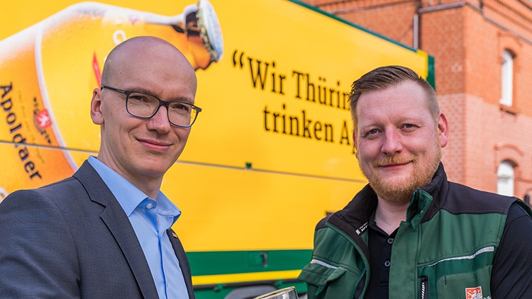 Zwei Personen stoßen mit Glasbier an, im Hintergrund ist ein gelber Lastwagen mit der Aufschrift „Apoldaer Glocken Hell“.