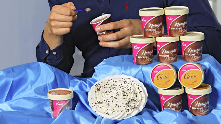 Person in dunkelblauem Hemd hält einen Becher Viba Nougat Eiscreme, weitere Eisbecher und ein Teller mit Streuseln stehen auf blauem Stoff.